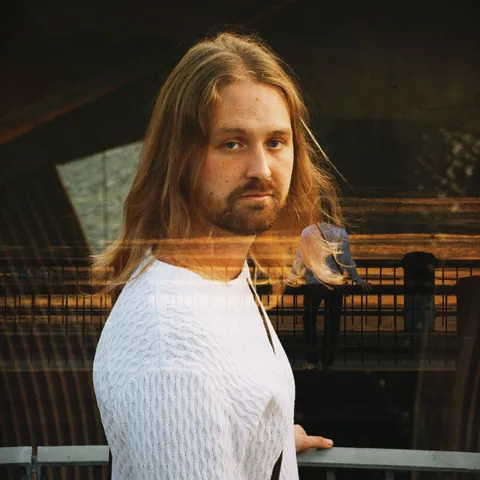 Tobias Lund in a white knit sweater, looking back, a double exposure over a railing in warm light